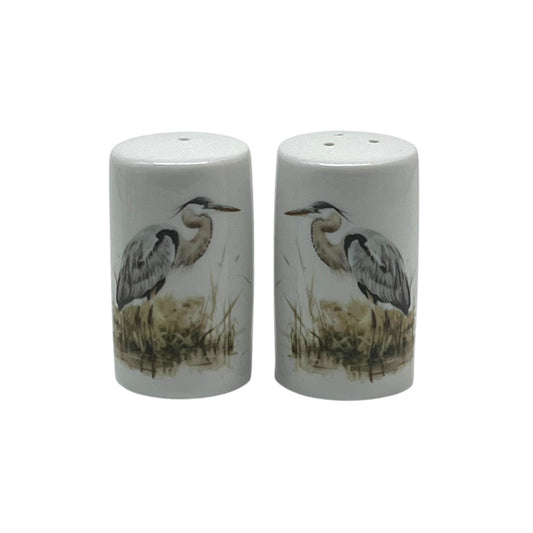 Pair of cylindrical white porcelain salt and pepper shakers featuring blue herons standing among softly painted marsh reeds.